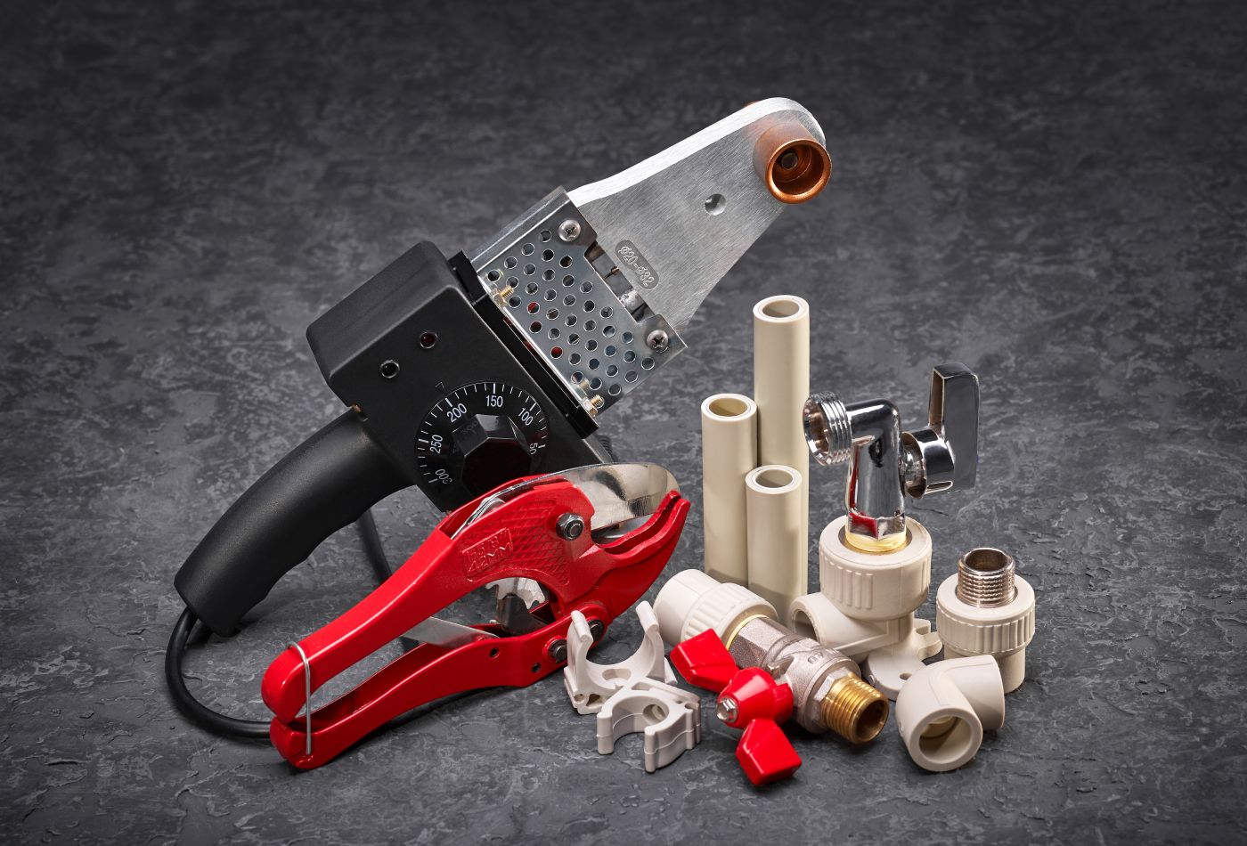  Assorted plumbing tools and materials on a dark surface, including a pipe welding machine, red pipe cutter, fittings, and plastic pipes.